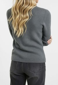 Donna bionda con capelli mossi, che indossa un maglione grigio aderente e jeans neri, in piedi con le braccia incrociate, voltata di spalle rispetto alla telecamera.