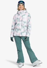Ski jacket in pastel pink and green with tree print, featuring a high collar and zip pockets. Paired with teal ski pants and white ski boots.
