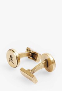 Gemelos de cuello de latón con un disco redondo que presenta un diseño de calavera y un cierre rectangular, acabado pulido y un brazo de conexión curvado.