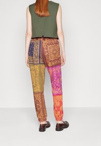 Top verde corto acompañado de pantalones multicolores y estampados que presentan parches amarillos, morados y rojos, con puños elásticos y zapatos marrones.