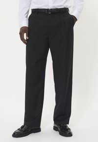 Czarne spersonalizowane spodnie z plisowanym przodem, wykonane z gładkiego materiału. W połączeniu z błyszczącymi czarnymi butami o fakturowanym wzorze.