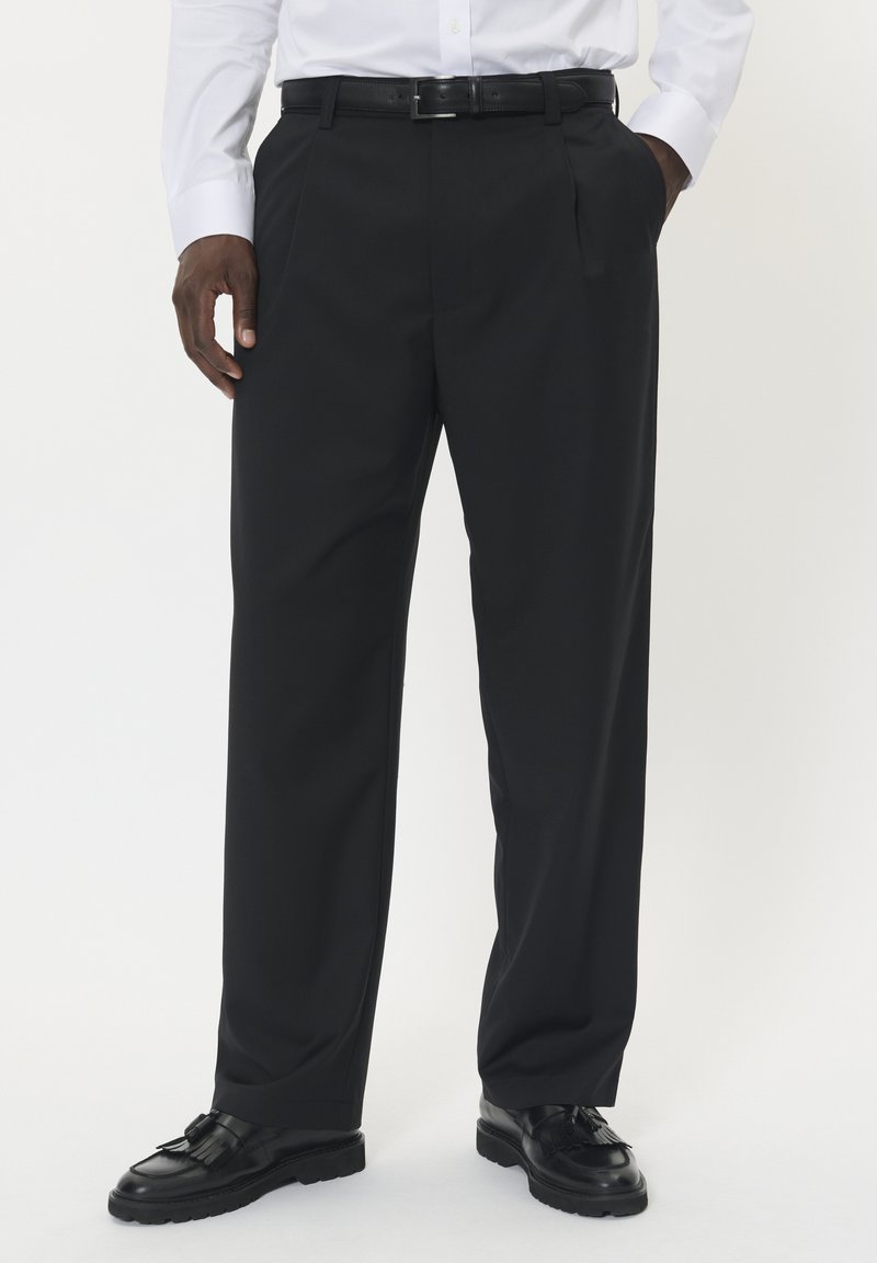 Czarne spersonalizowane spodnie z plisowanym przodem, wykonane z gładkiego materiału. W połączeniu z błyszczącymi czarnymi butami o fakturowanym wzorze.