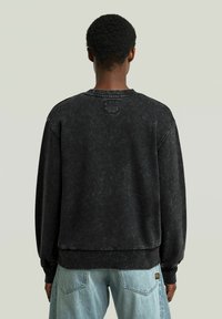 Felpa nera con collo rotondo, caratterizzata da una texture sbiadita e una leggera usura. Maniche a costine e orlo a coste, con una piccola toppa sulla parte posteriore del colletto.