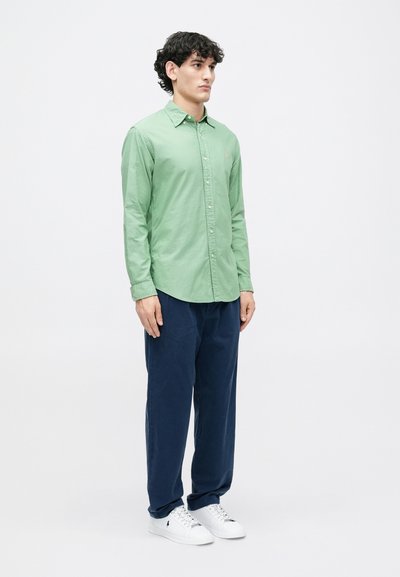 Jovem em pé, vestindo uma camisa de botão verde-claro, calças azul-marinho e ténis brancos, contra um fundo branco simples.