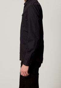 Camicia nera a maniche lunghe con colletto a bottone, dotata di taschino frontale e taglio dritto. Dettagli delle cuciture e polsini in vista. Materiale in cotone.