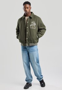 Olive green wool blend jacket with a large embroidered letter detail, paired with light blue baggy jeans and black sneakers.