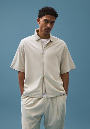 Bărbat cu păr scurt și creț purtând o cămașă bej cu mânecă scurtă cu fermoar peste un tricou alb și pantaloni bej asortați pe un fundal albastru.