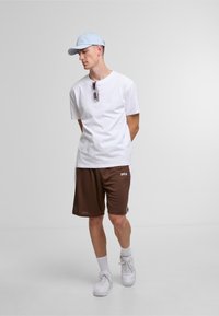 Chemise blanche à manches courtes, shorts marron avec logo, casquette bleu clair et tennis blanches. Des lunettes de soleil reposent sur la chemise. Tenue simple et décontractée.