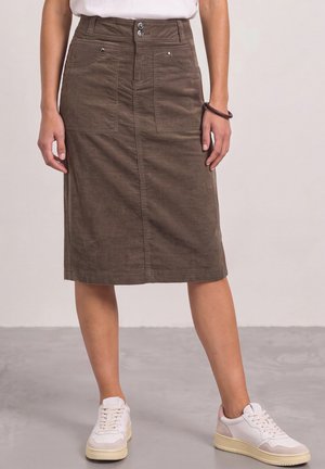Kvinde iført brun knee-length kordnert nederdel med forlommer og hvide sneakers, stående på en grå gulv mod en ensfarvet baggrund.