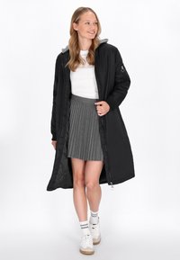 Abrigo largo negro con capucha gris, con cierre de cremallera, textura acolchada y bolsillos laterales, llevado sobre una camisa blanca y una falda gris con pliegues.