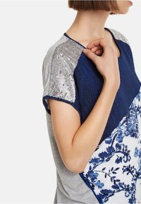 Top de manga corta con un diseño floral en azul marino y blanco, hombros acentuados con lentejuelas plateadas y espalda gris. Tejido de múltiples texturas.