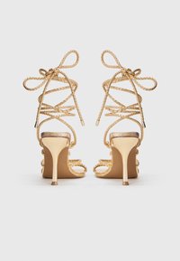 Sandalias de tacón alto doradas con tiras trenzadas, punta abierta y un tacón delgado, con una textura suave y un diseño de sujeción al tobillo seguro.