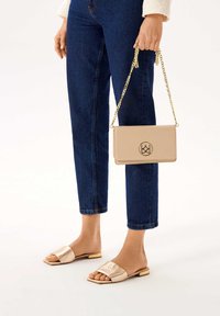 Kahová kožená crossbody taška so zlatým reťazovým popruhom, s detailom loga na prednej strane. Model má na sebe tmavomodré džínsy a svetlo zlaté páskové sandále.