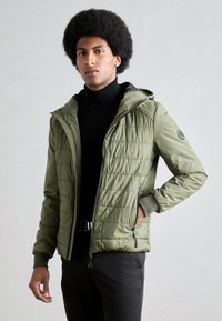Veste matelassée vert olive avec capuche amovible, fermeture éclair sur le devant et poches latérales. Présente un matelassage horizontal et des poignets côtelés.