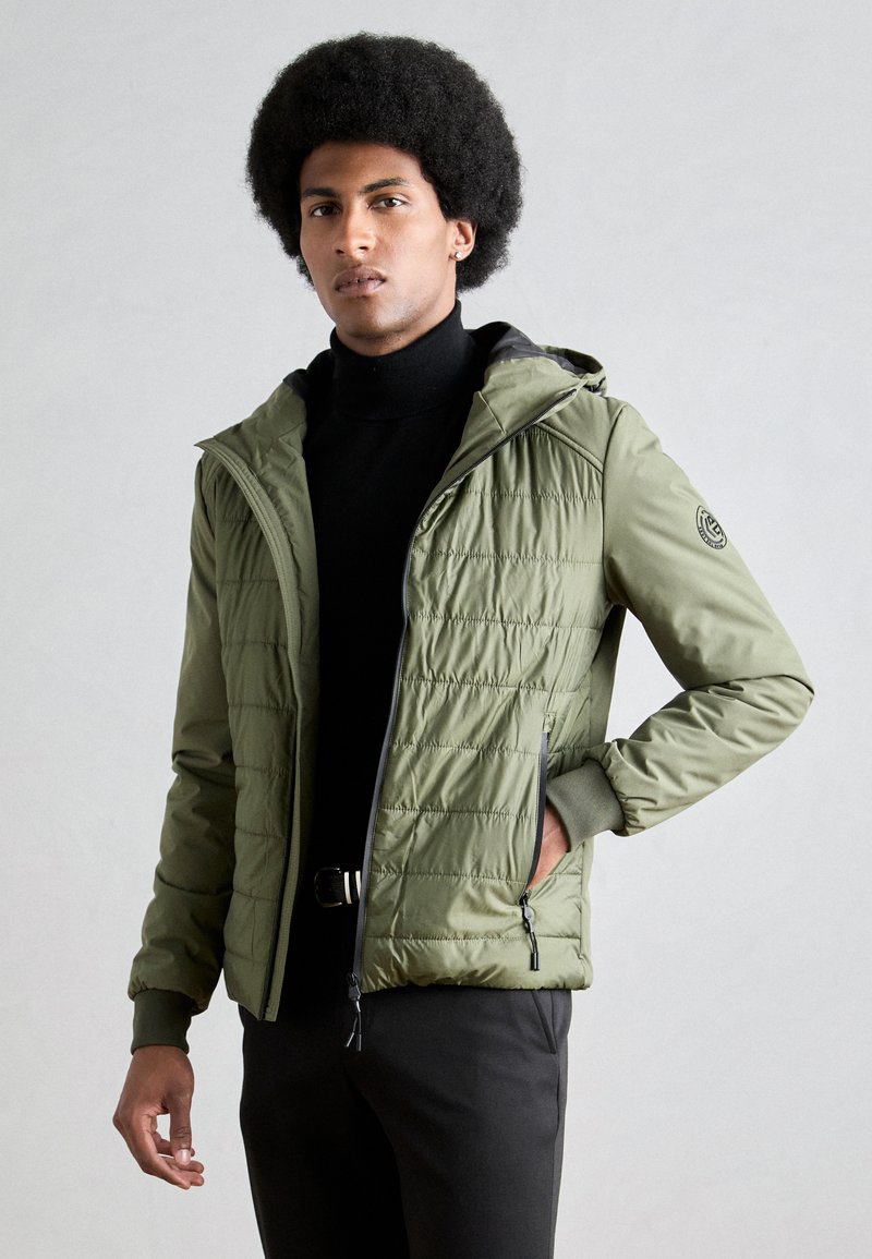 Veste matelassée vert olive avec capuche amovible, fermeture éclair sur le devant et poches latérales. Présente un matelassage horizontal et des poignets côtelés.