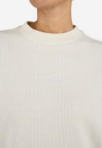 Maglione a coste beige chiaro con collo rotondo. Presenta il logo ricamato "PEGADOR" in blu chiaro sul davanti. Aspetto della stoffa texture.