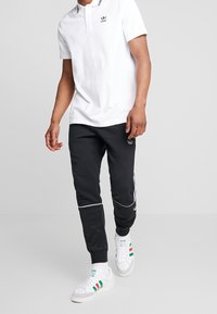 En person som bär en vit Adidas-polo, svarta träningsbyxor med vita ränder och vita sneakers med gröna och röda accenter går framåt.