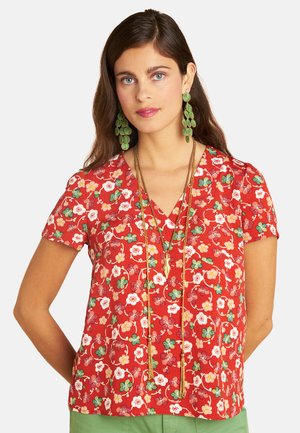 Woman with long brown hair wears a red floral top, long green earrings, layered gold necklaces, and green pants, standing against a plain background.