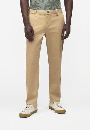 Muž v béžových nohaviciach rovného strihu, bielych teniskách a zelenej košeli s potlačou palmových listov, stojaci pred jednoduchým pozadím.