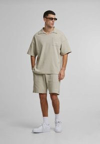 Beige kortermet polo med lomme og matchende shorts, begge i et teksturert stoff. Brukt med hvite joggesko og brune solbriller.