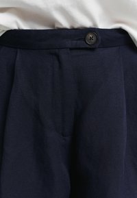 Pantalons plissés bleu marine en tissu texturé, dotés d'une fermeture à bouton visible et d'un détail de poche latérale.