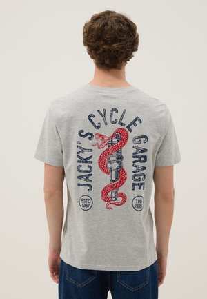 Man met krullend haar in grijs T-shirt toont een rode slang die om een bougie is opgerold en tekst "Jacky's Cycle Garage" op de achterkant.