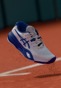 Baskets blanches avec des accents bleus, tige en maille, semelle texturée et lacets visibles, présentées sur un court en terre battue avec des particules de terre.