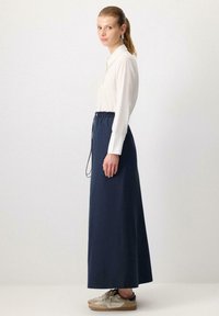Long navy skirt with an elastic waistband and drawstring detail, paired with a light-colored button-up shirt and beige sneakers.