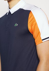 Marinblå polo skjorta med orange och vita ärmar, tillverkad av slätt tyg, med en liten grön krokodil-logotyp på bröstet.