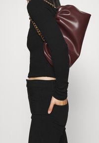 Burgundy leather tote bag with a slouched design and a gold chain strap, paired with a black fitted long-sleeve top and black pants.