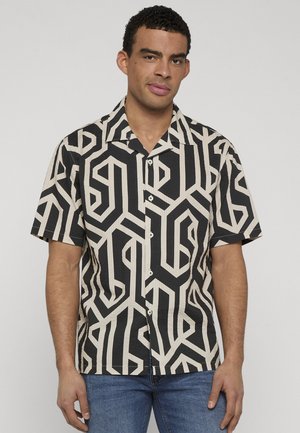 Mand iført en sort og beige kortærmet skjorte med geometrisk mønster og blå jeans, der står mod en ensfarvet lys baggrund.