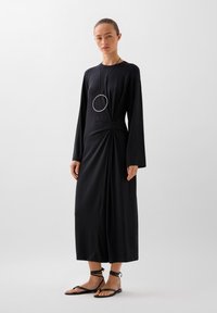 Abito lungo nero con dettaglio a nodo in vita. Realizzato in tessuto liscio, presenta una silhouette fino al pavimento e maniche slanciate.