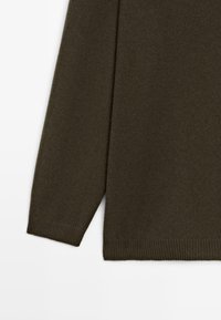 Manica e orlo inferiore di un maglione lavorato a maglia verde oliva scuro con polsini e vita a costine su sfondo bianco.