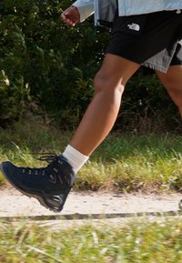 Black hiking boots with high ankle support, laces, and rubber sole, worn with white socks. Background features grass and a dirt path.