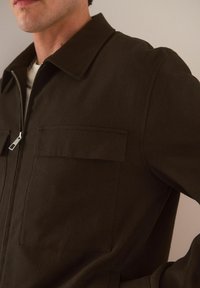 Chaqueta de cremallera verde oscuro hecha de un tejido texturizado, con un cuello puntiagudo y dos bolsillos delanteros con solapas rectangulares.