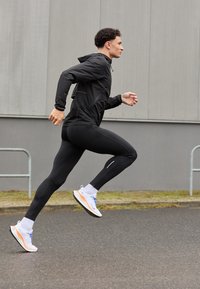 Mann in schwarzer Sportbekleidung, der auf einer gepflasterten Straße neben einem grauen Gebäude läuft, im mittleren Lauf mit einem Fuß in der Luft.