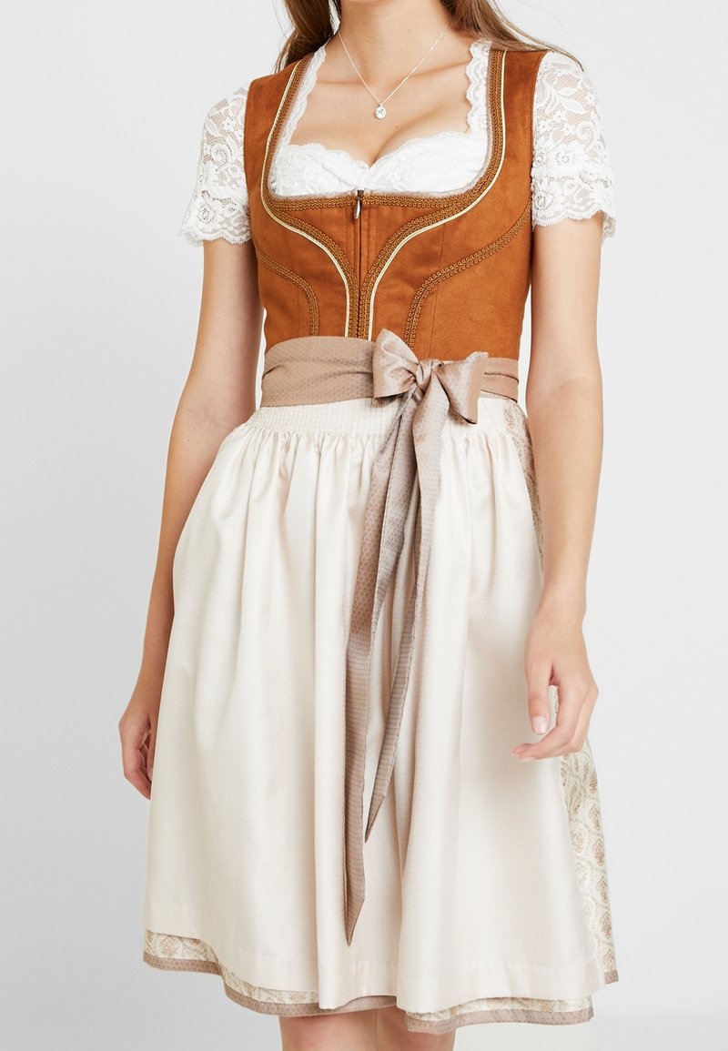 Femme portant un chemisier en dentelle blanche sous un corsage en daim marron et une jupe crème avec une ceinture beige nouée à la taille.