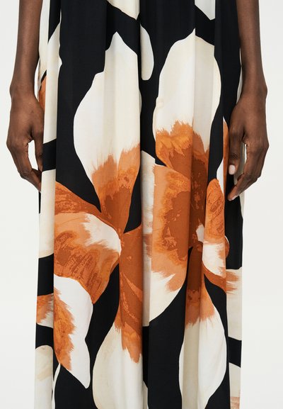 Τα χέρια ξεκουράζονται στα πλευρά, φοράνε ένα ένδυμα με μοτίβο από μαύρα, λευκά και πορτοκαλιά άνθη με μεγάλα πέταλα.