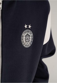 Navy and white jacket sleeve with zipper and embroidered "Prohibited Berlin" crest featuring two stars above it.