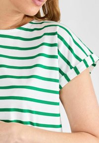 Camiseta blanca con rayas verdes horizontales, con mangas cortas enrolladas y escote redondo. Material de algodón suave con una textura lisa.