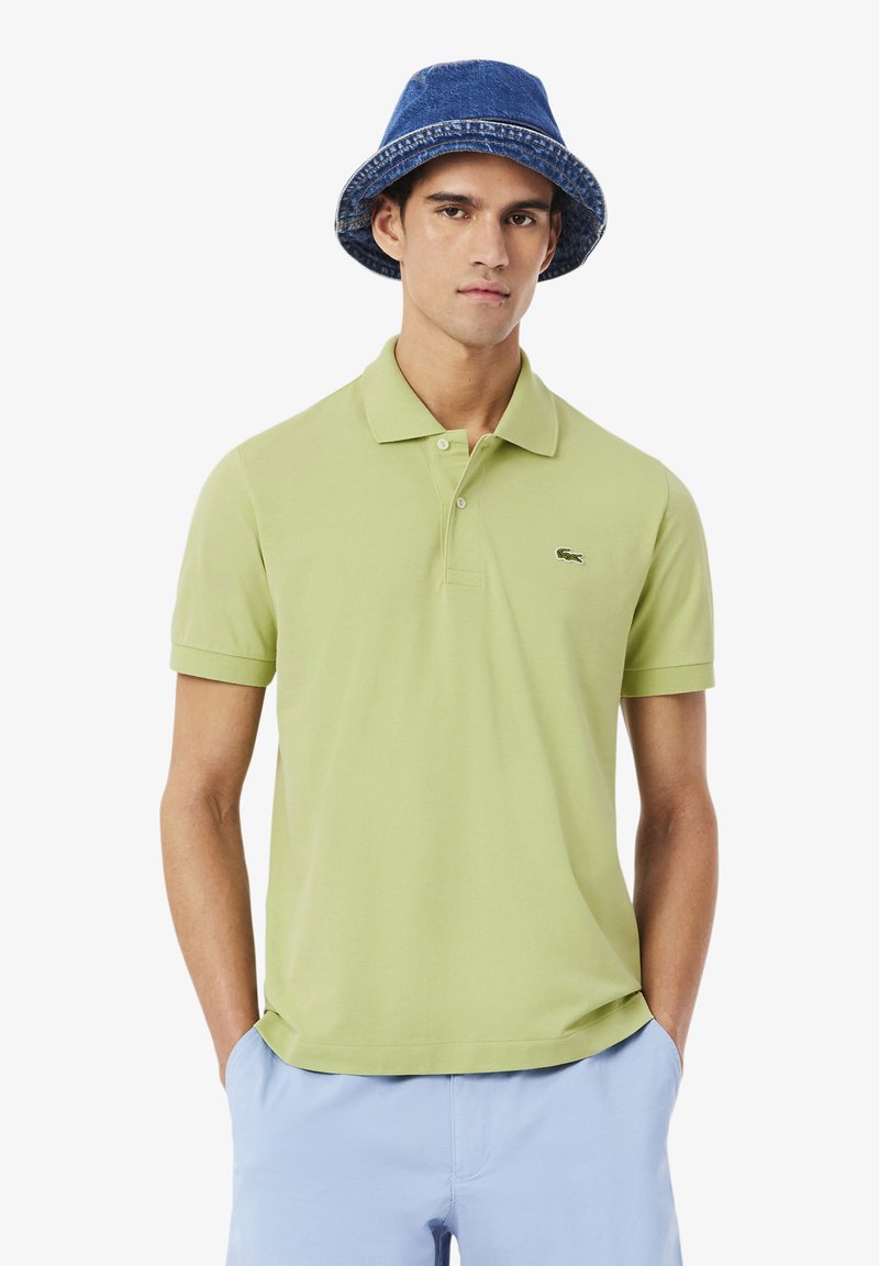 Jonge man die een lichtgroen poloshirt, lichtblauwe broek en een blauwe denim bucket hat draagt, staand met handen in zijn zakken.