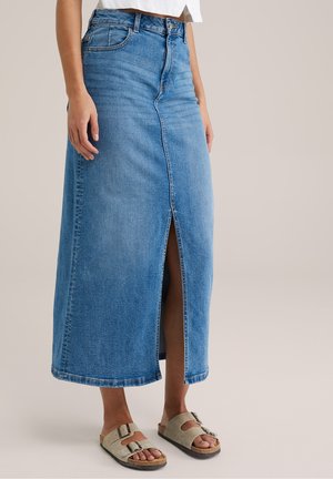Frau trägt einen blauen, hoch taillierten Denim-Midirock mit Frontschlitz, kombiniert mit beigen Sandalen mit Schnalle und einem weißen Cropped Top.