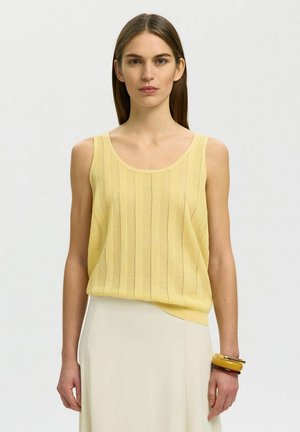 Kvinde med glat brunt hår iført gul ærmeløs striktop, cremefarvet nederdel og tre chunky armbånd på højre håndled, står mod hvid baggrund.