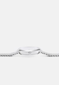 Montre en argent avec un boîtier rond, un bracelet en métal maillé et un design minimaliste. Elle dispose d'une surface en verre lisse et d'une couronne latérale.