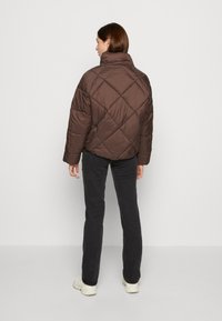 Veste en duvet matelassée marron avec un col montant, présentant une texture brillante et un motif de couture diagonal. Portée sur un pantalon noir.