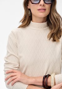 Vrouw in een gestructureerde beige coltrui, met schildpadprint zonnebril, een decoratieve oorbel en drie armbanden op gekruiste armen.