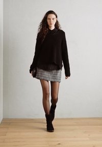 Czarny sweter z dzianiny o luźnym kroju, w połączeniu z mini spódnicą w szaro-czarny wzór oraz czarnymi kozakami do kolan. Gładka drewniana podłoga.