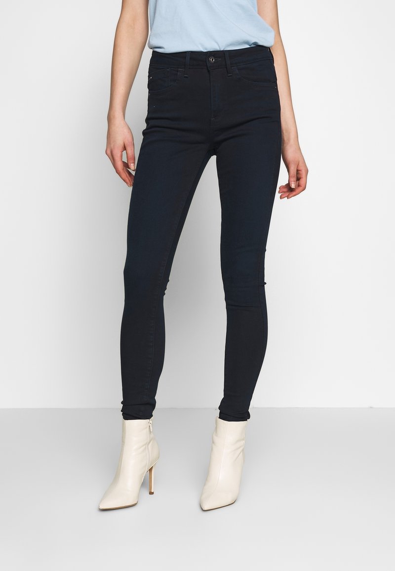 Jeans skinny de un azul oscuro con una cintura alta, que presentan bolsillos frontales y detalles de costura sutiles, combinados con botas de tobillo de tacón en color crema.