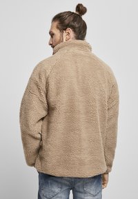 Beige fleecejakke med høj krave, struktureret overflade og raglanærmer. Båret over blå denim jeans, set fra ryggen.
