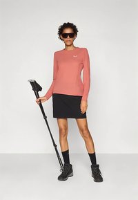 Nő napszemüveget visel, korall színű hosszú ujjú polót, fekete szoknyát és túracipőt hord, kezében egy összecsukott túrabotot tart, egy egyszínű háttér előtt áll.